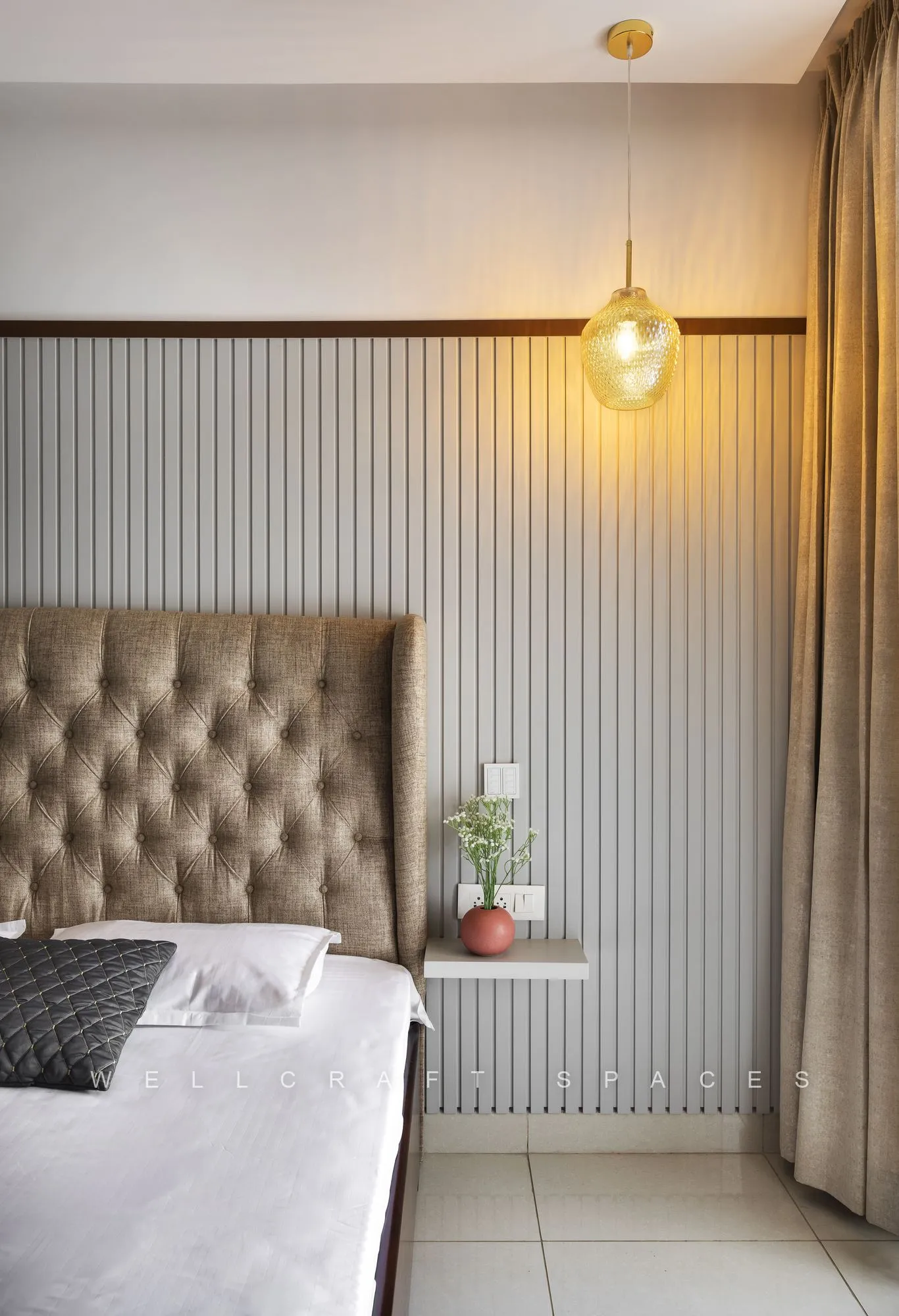 Tufted bedhead against a ribbed cream wall, lit by a hanging amber glass pendant beside a small planted ledge.