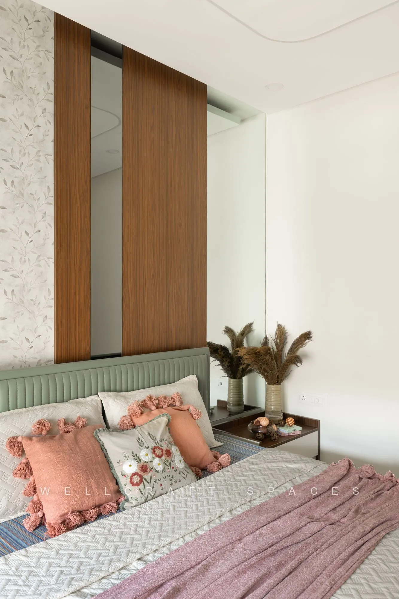 Bed with a sage-green channelled headboard, pink and floral cushions and a teak-framed textured panel above the window seat.
