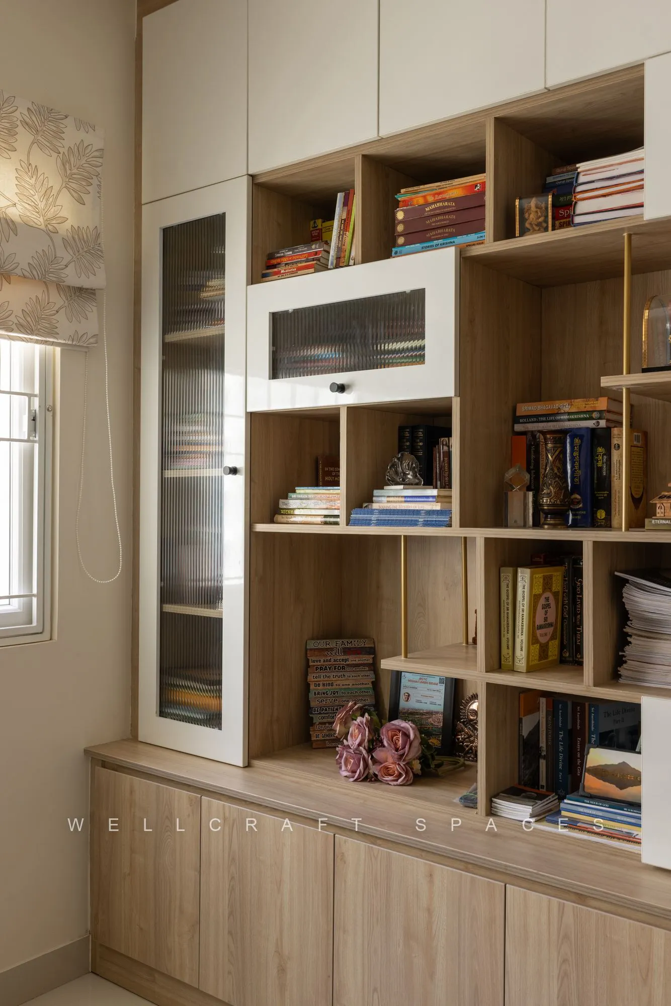 Floor-to-ceiling oak bookshelf packed with books and objects above a closed lower cabinet run, beside a shuttered window.