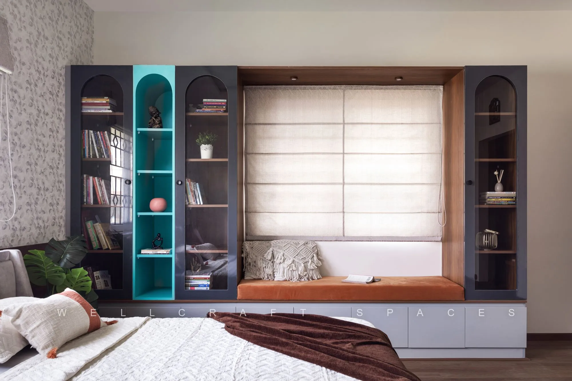 Built-in wall of walnut and teal display cabinets flanking a cushioned window seat, with arched glazed shutters and books and ceramics on the shelves.