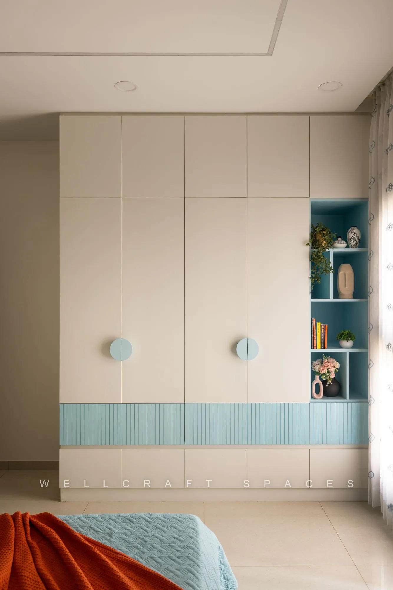 Cream wardrobe with a sky-blue fluted band and round handles, flanked by a turquoise open bookshelf.