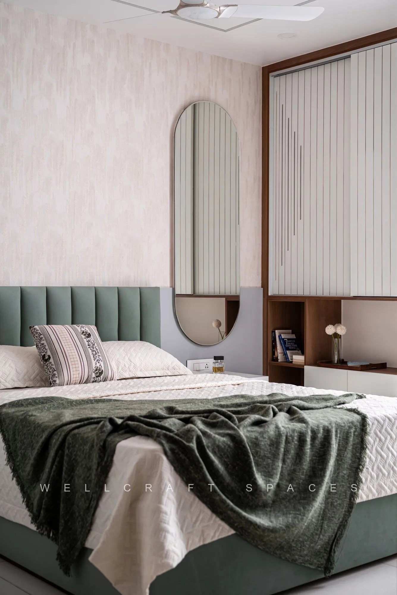 Bedroom in sage and oatmeal with a green channel-tufted headboard, an oval brass mirror, a moss-green throw and vertical blinds at the window.