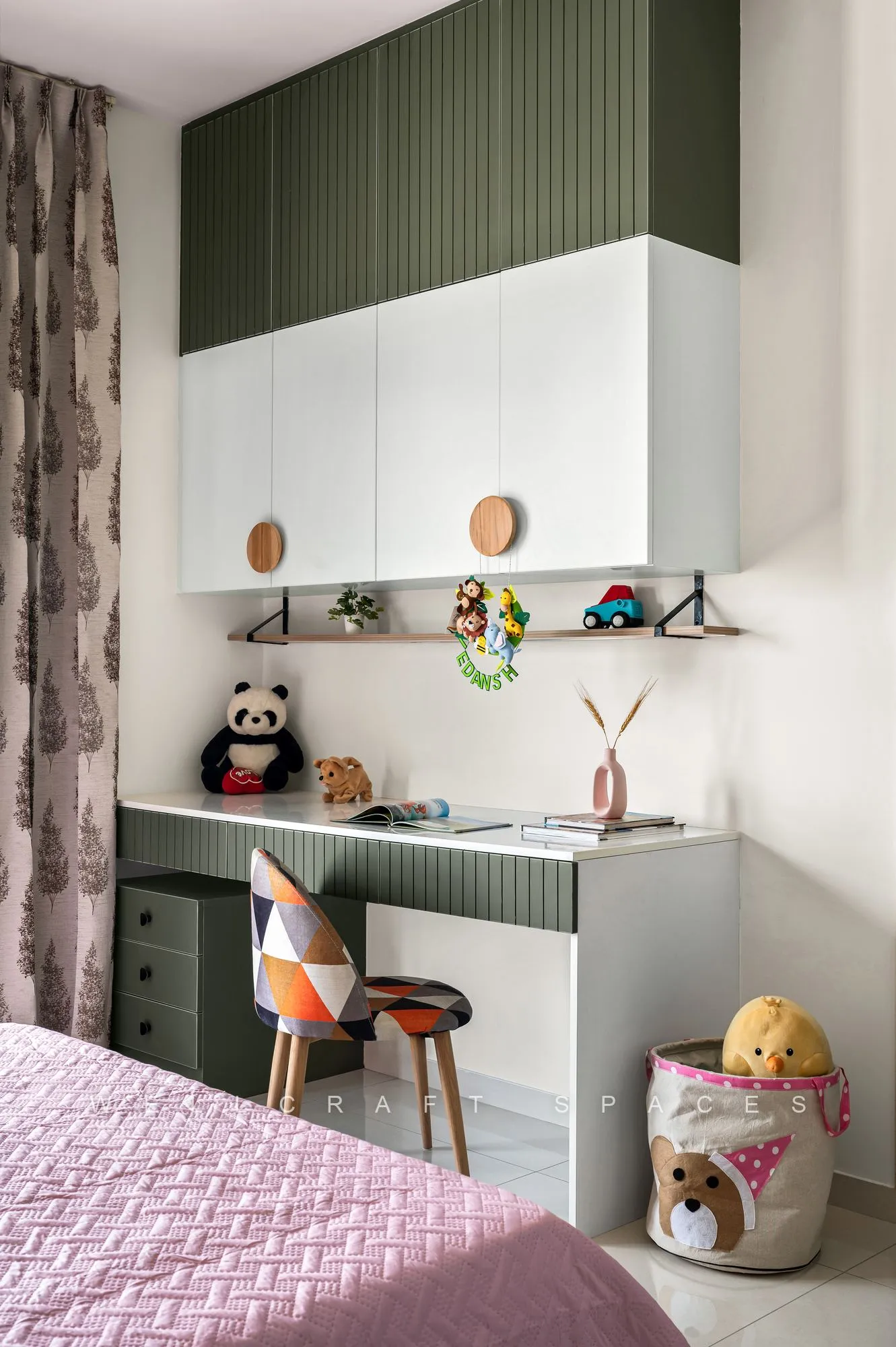 Study corner in the child's room with a sage-green fluted overhead unit, a compact desk, soft toys and a patterned stool.