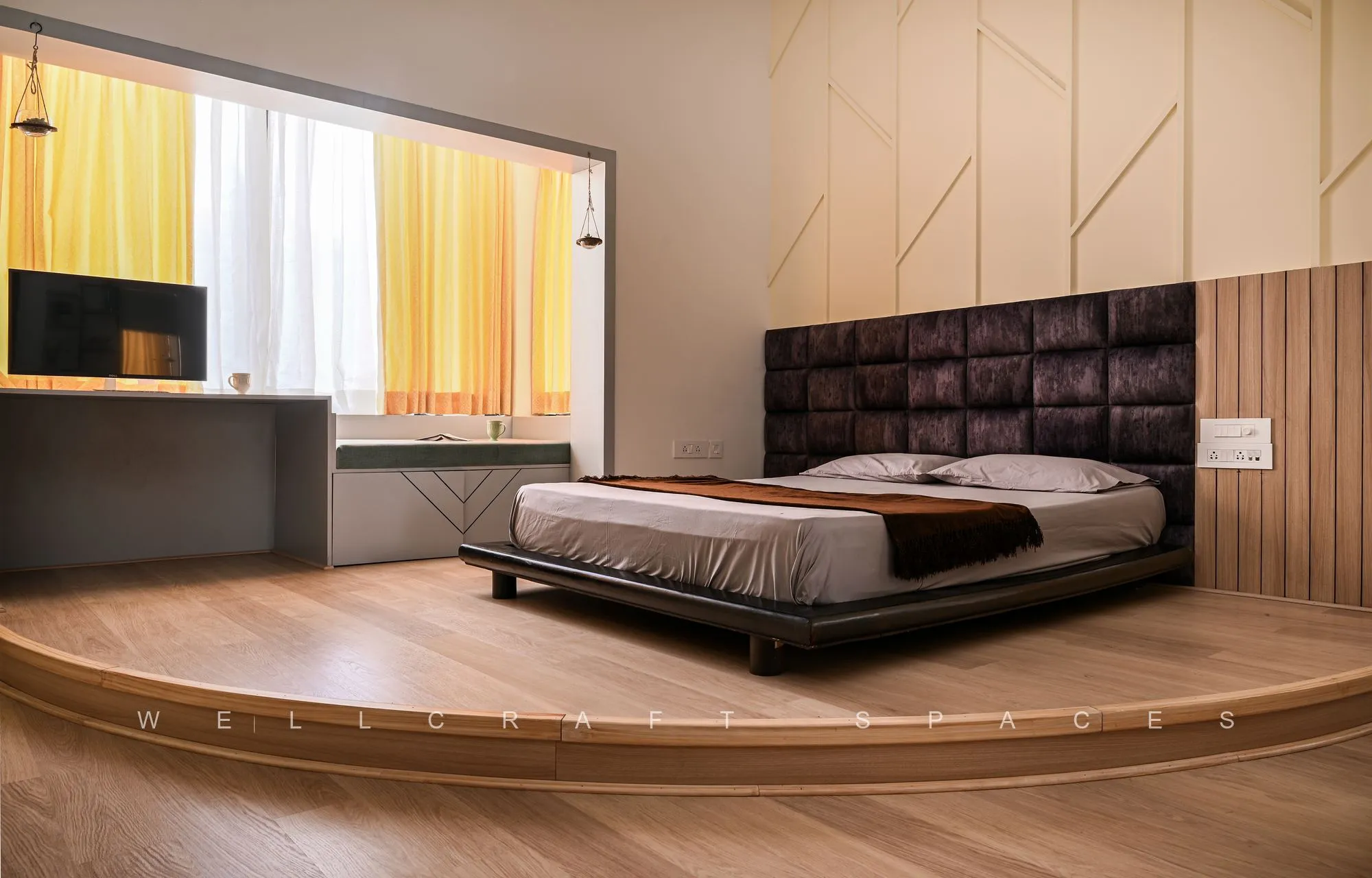 Master bedroom on a curved raised timber platform, with a quilted charcoal leather headboard, cream fluted panelling and a long yellow-lit window ledge.