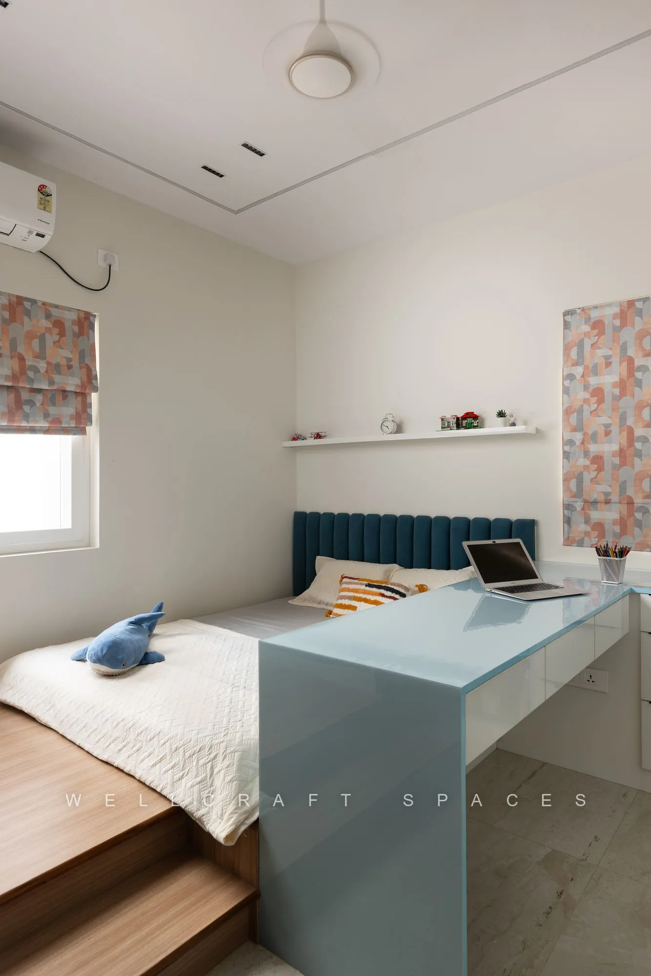 Child's room with a raised timber platform bed, a duck-egg blue desk counter, floating shelves and a patterned roman blind.