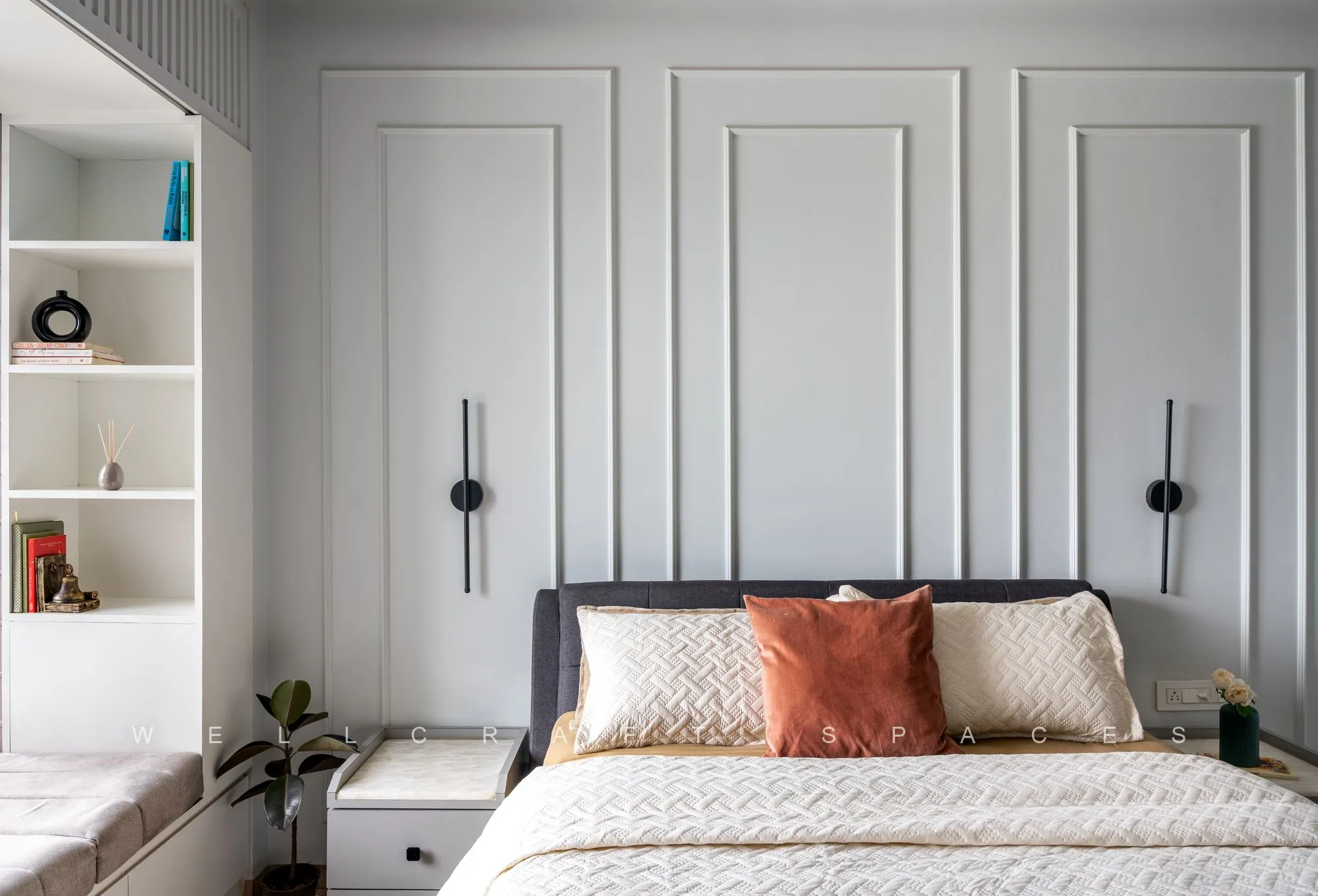 Bedroom with a grey moulded panel headboard wall, rust and cream cushions, black wall sconces and built-in niches holding books and greenery.