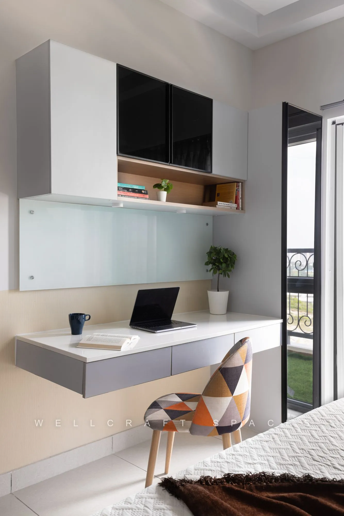Wall-mounted white study desk with a laptop, frosted glass overhead cabinets and a geometric-print chair beside the balcony door.
