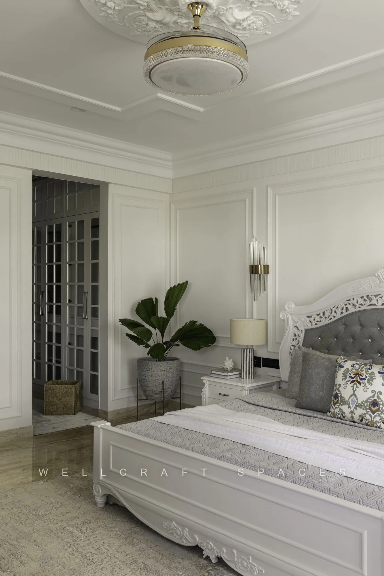 Guest bedroom in classic white with a carved four-post bed, grey buttoned headboard, glass flush ceiling light and a potted palm by the doorway.