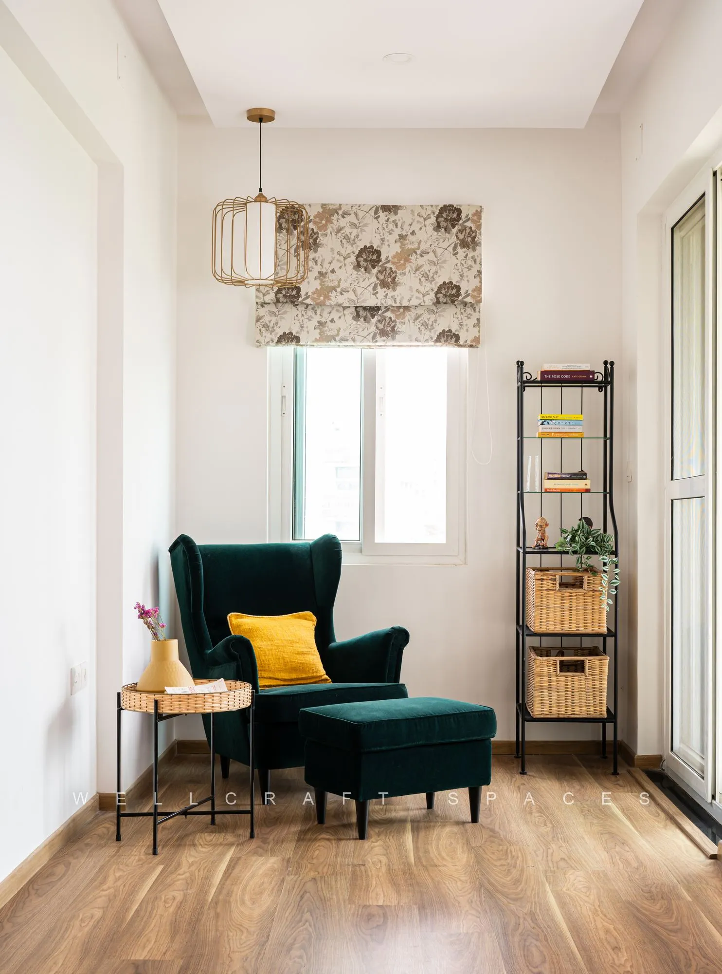 Reading corner with a bottle-green wing chair and matching ottoman, a mustard cushion, a black metal etagere and a floral roman blind.