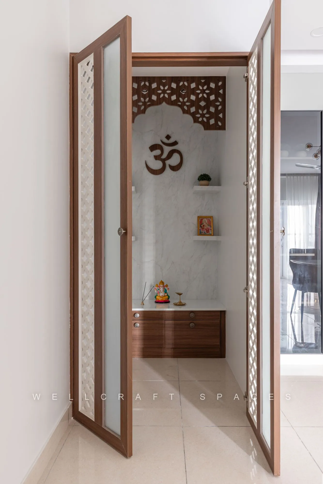 Pooja room behind carved teak-framed doors with an Om-etched fretwork screen, a wooden mandap and shelf for lamps.