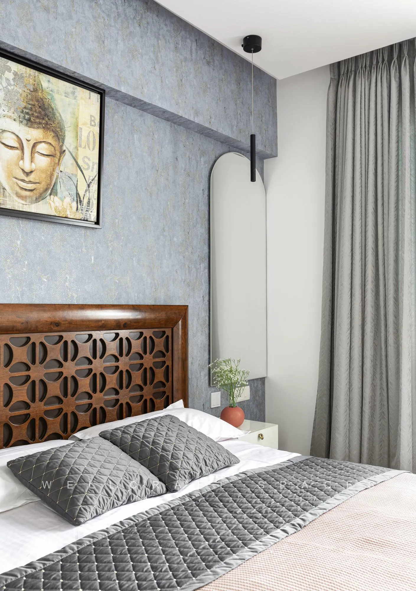 Bed with a carved lattice teak headboard against a pale blue textured wall hung with a Buddha painting.