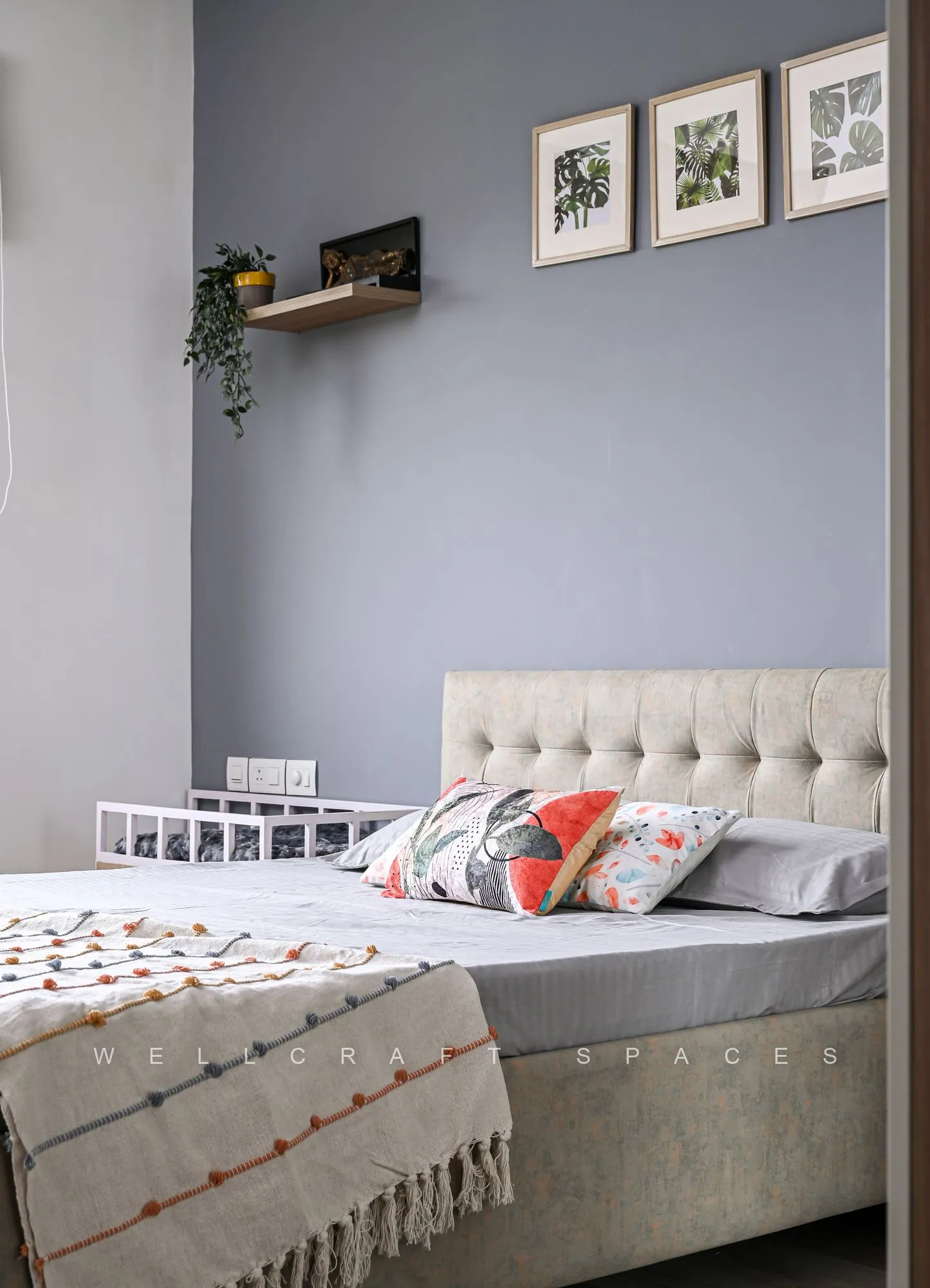 Bedroom with a buttoned cream headboard against a grey wall, framed botanical prints above and a woven ivory bedcover.