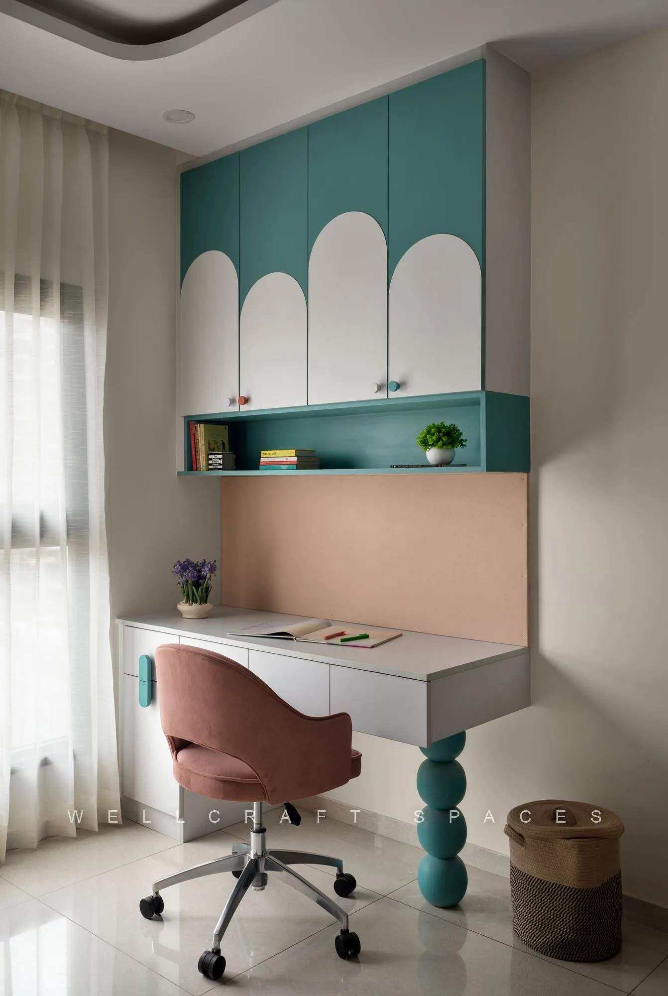 Study corner with a white desk under a teal overhead cabinet, arched wall graphics, open shelves and a blush swivel chair.