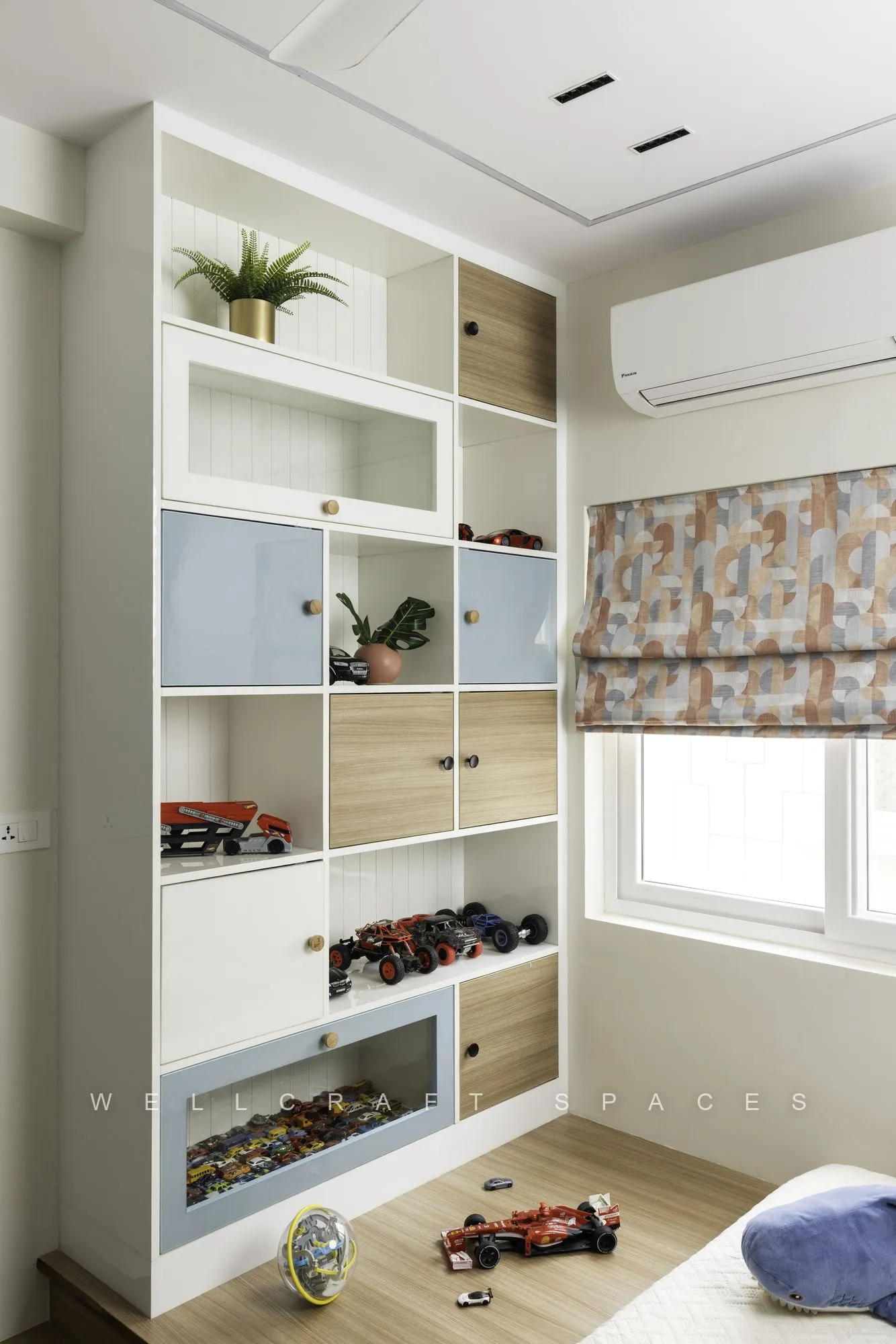 Wall of white and pale blue open shelving filled with toy cars and models, beside a window with a printed blind and toys on the floor.