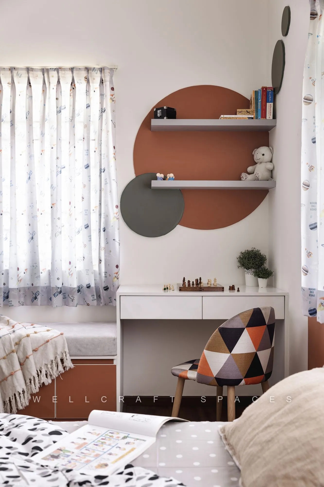 Child's room with overlapping terracotta and olive circles painted behind floating shelves, a white study desk and a triangle-patterned chair by the window.