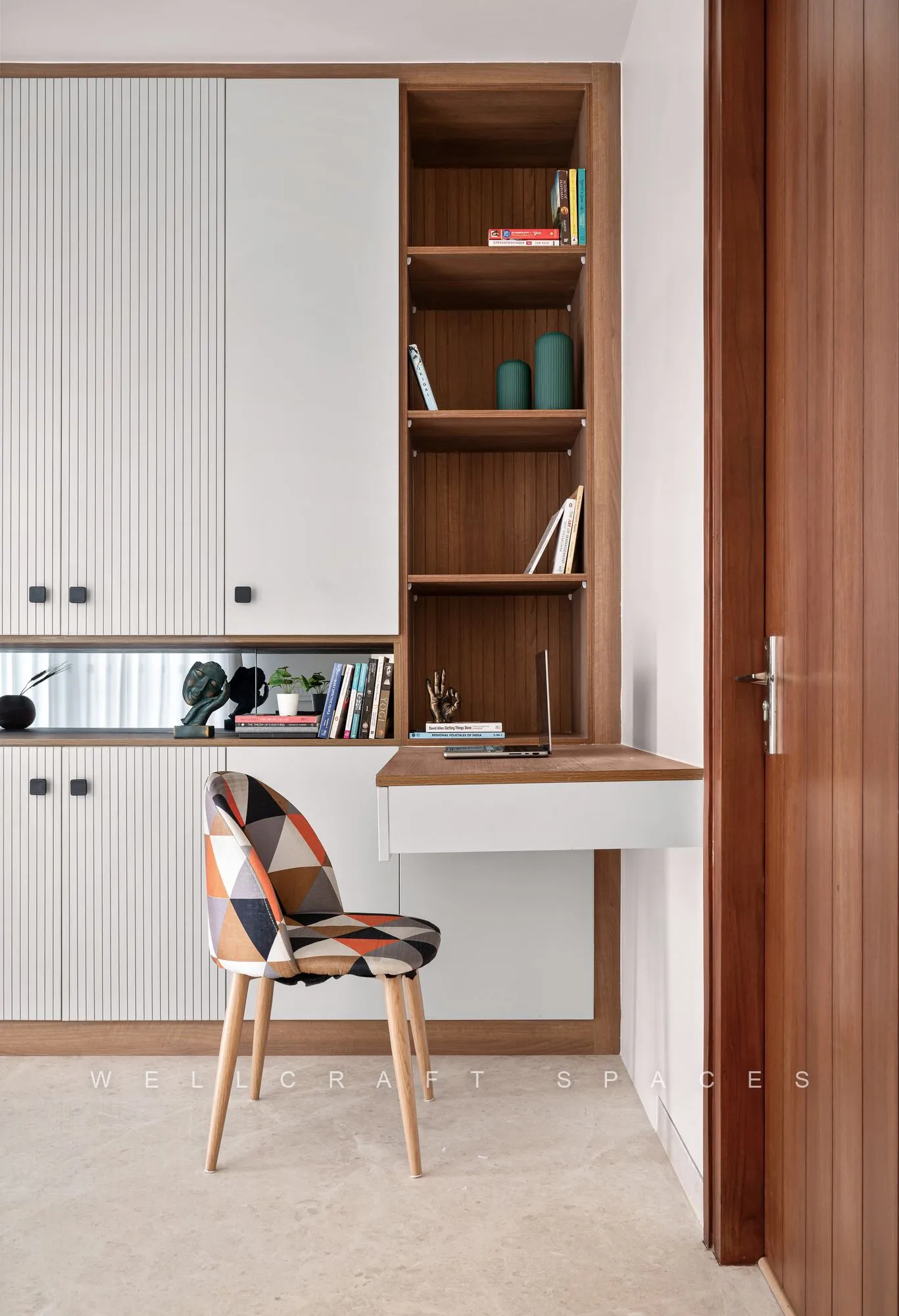Study nook with a white fluted cabinet run, walnut open shelving, a slim desk and a patchwork-print chair on pale timber legs.