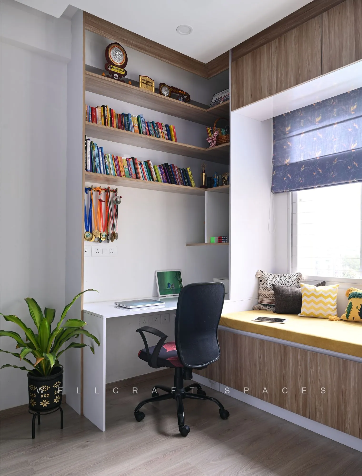 Study with a white corner desk, black task chair, wall-mounted bookshelves and a cushioned window seat under a roman blind.