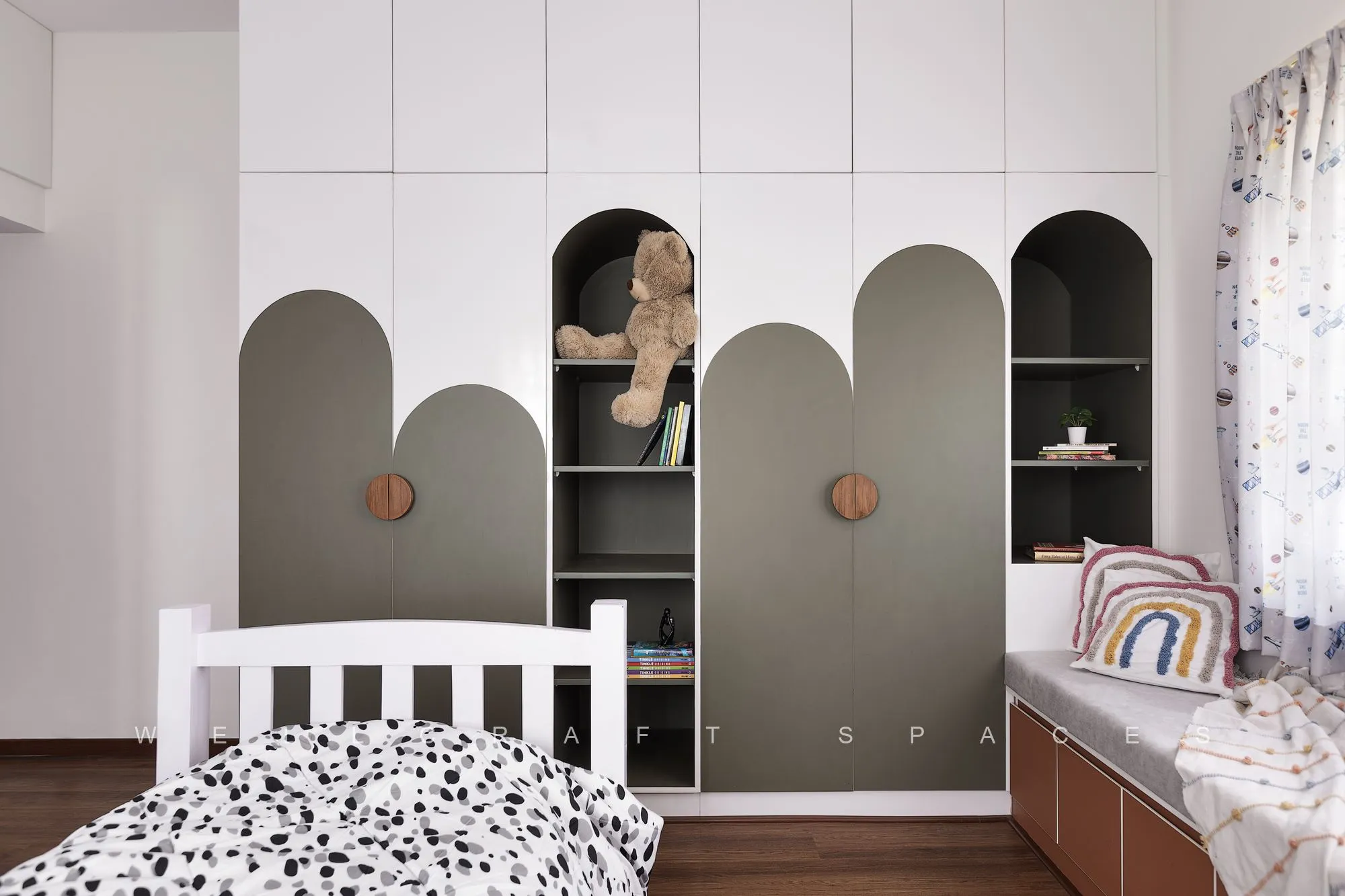 Kids' room with arched olive and white wardrobe shutters and open niches for toys, a white spindle bed with a black dot-print quilt and a window bench.