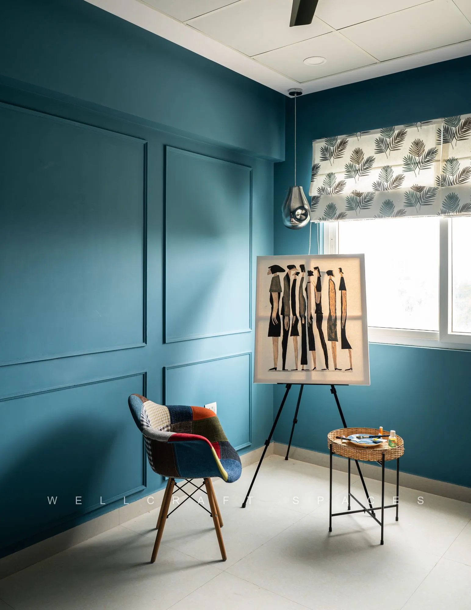 Work corner against a deep teal panelled wall, with an artwork on a black easel, a moulded chair on timber legs and a small black side table.