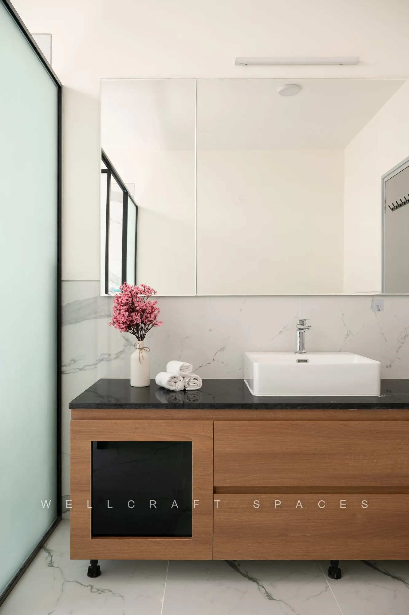 Bathroom vanity in walnut with a black stone counter, a white vessel basin, a full-width frameless mirror and a fluted glass partition.