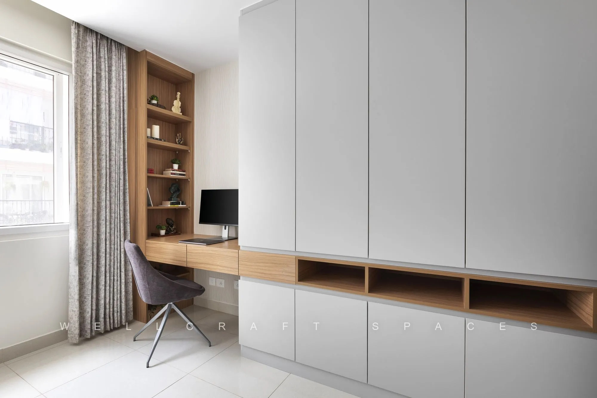 Built-in study with a teak desk and open shelving between tall grey wardrobes, a swivel chair and a monitor by the window.