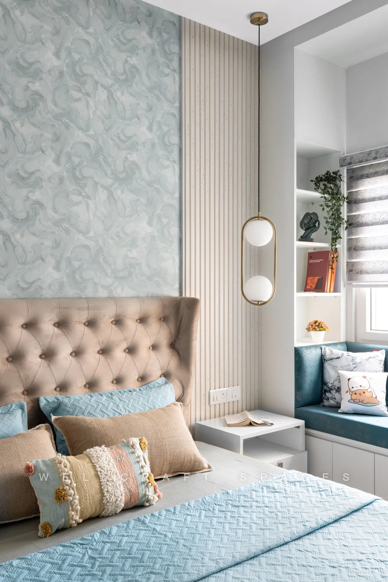 Bedroom with a tufted beige velvet headboard against pale blue marbled wallpaper, a brass pendant with glass globes, and a teal window-seat nook lined with bookshelves.