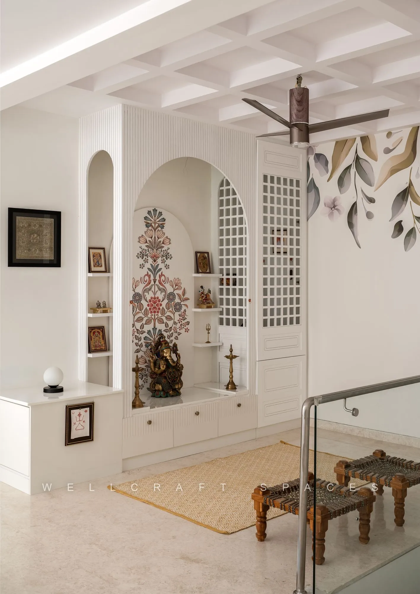 Pooja alcove in an arched white niche with a carved marble idol, jali screen doors, a low wooden altar and painted leaf motifs on the adjoining wall.
