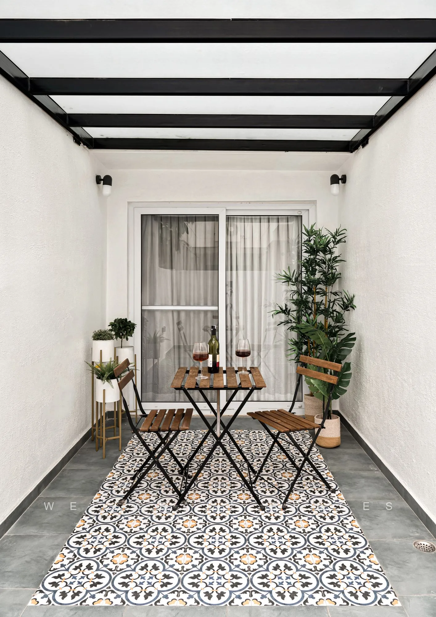 Narrow open terrace laid with black-and-white patterned encaustic tiles, a folding bistro table and chairs, and potted plants along a white parapet.