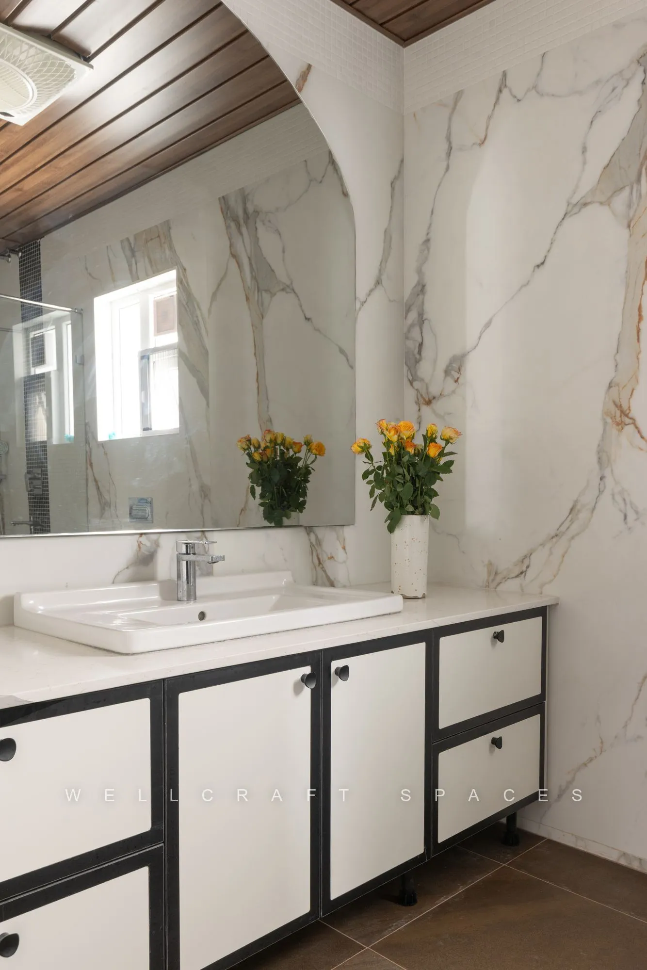 Marble bathroom with a white twin-drawer vanity, black framed handles, large mirror and a vase of yellow blooms.