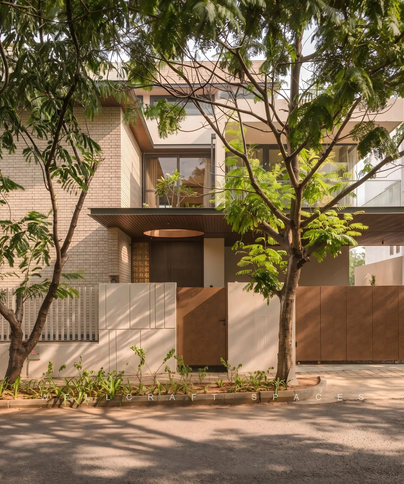 Street elevation of the brick and plaster villa seen through mature trees, with deep timber-framed openings and a low boundary wall.