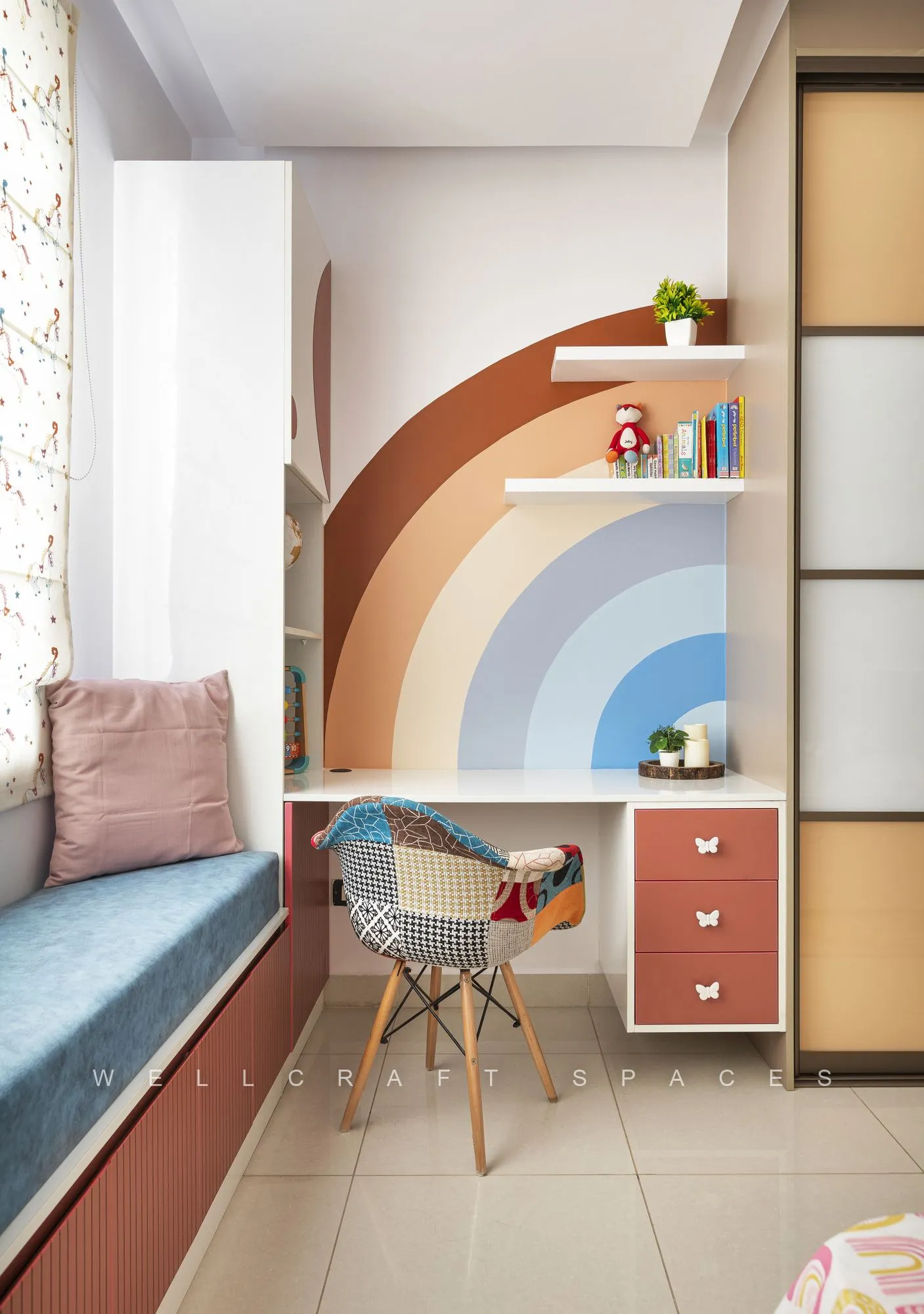 Kids' room with a hand-painted rainbow mural above a built-in study desk, terracotta drawers with butterfly pulls, a patchwork armchair and a blue window seat.